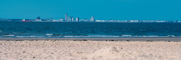 Panoramic view of an urban skyline across a calm blue sea, with a sandy beach in the foreground.