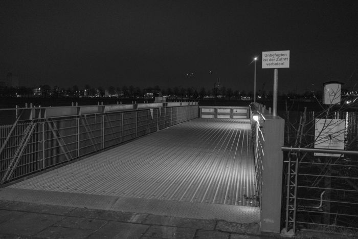 Empty bridge at night, illuminated by a single light, with a cityscape in the background and a sign reading 