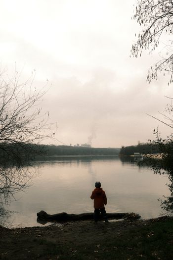 A person in an orange jacket stands by a calm lake at sunset, with bare trees framing the scene.