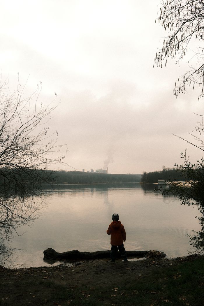 A person in an orange jacket stands by a calm lake at sunset, with bare trees framing the scene.