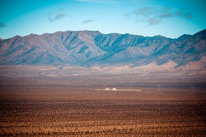 Mountains rise over vast desert landscape with a lone house in the distance, under clear blue sky.
