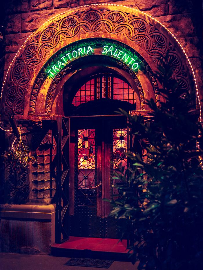 Restaurants entrance with ornate archway, neon sign, plants on either side, red carpet, and glass door.
