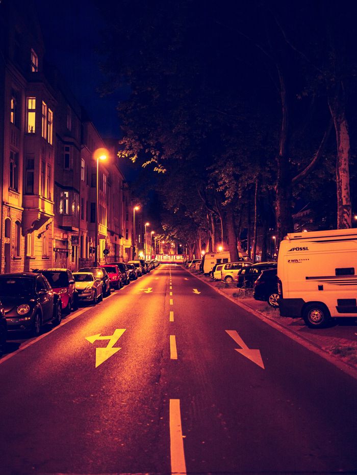 A dimly lit street at night with parked cars on both sides, trees lining the sidewalk, and arrows painted on the road.