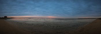 Sunset over calm ocean waters with a sandy beach in the foreground, under cloudy sky.
