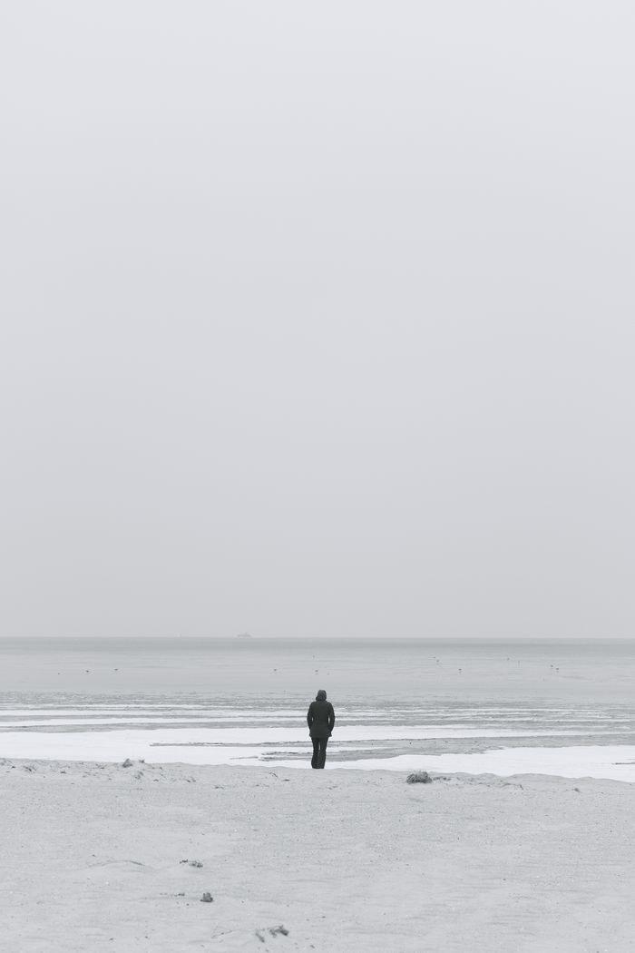 A solitary figure stands on a vast, empty beach with the ocean in the background.