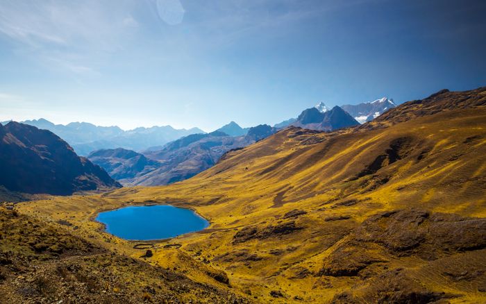 Mountainous landscape with a deep blue lake in the foreground. Golden hills roll towards snow-capped peaks under a bright sky.