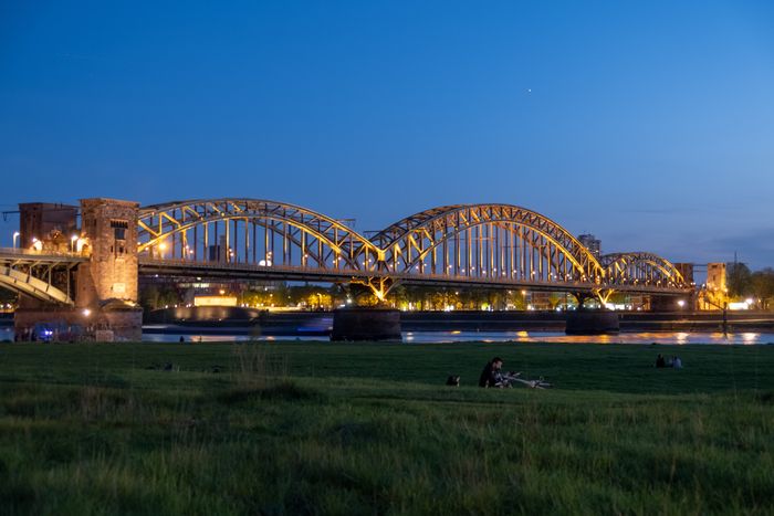 Lit bridge over river at dusk with people picnicking in foreground, grass, clear sky.
