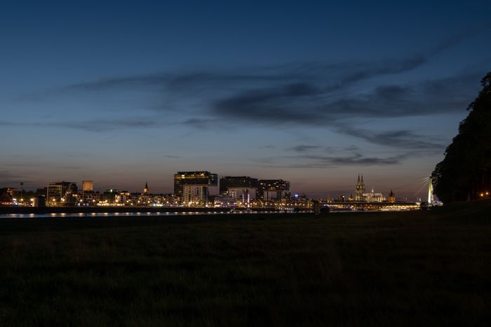City skyline at dusk, buildings illuminated against dark blue sky with scattered clouds, water in foreground.