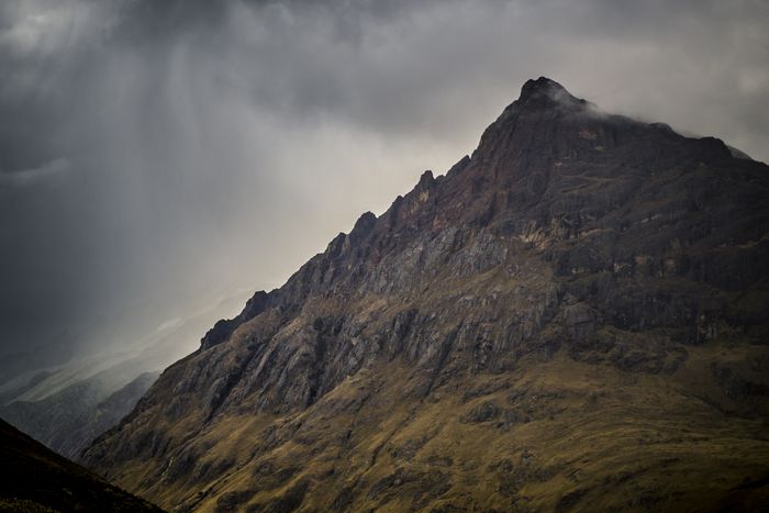 Dark, moody mountain peak with rocky face, set against dramatic cloud-filled sky.