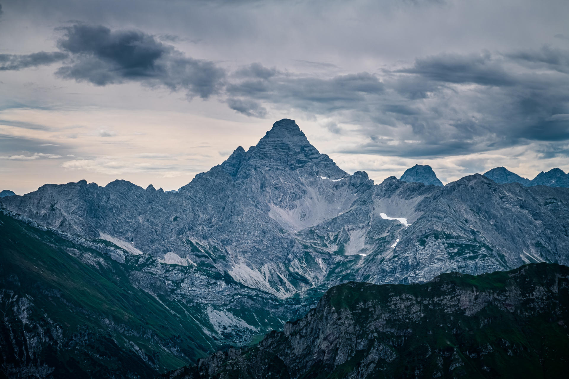 Majestic mountain peak under a cloudy sky, with rugged slopes and patches of snow.