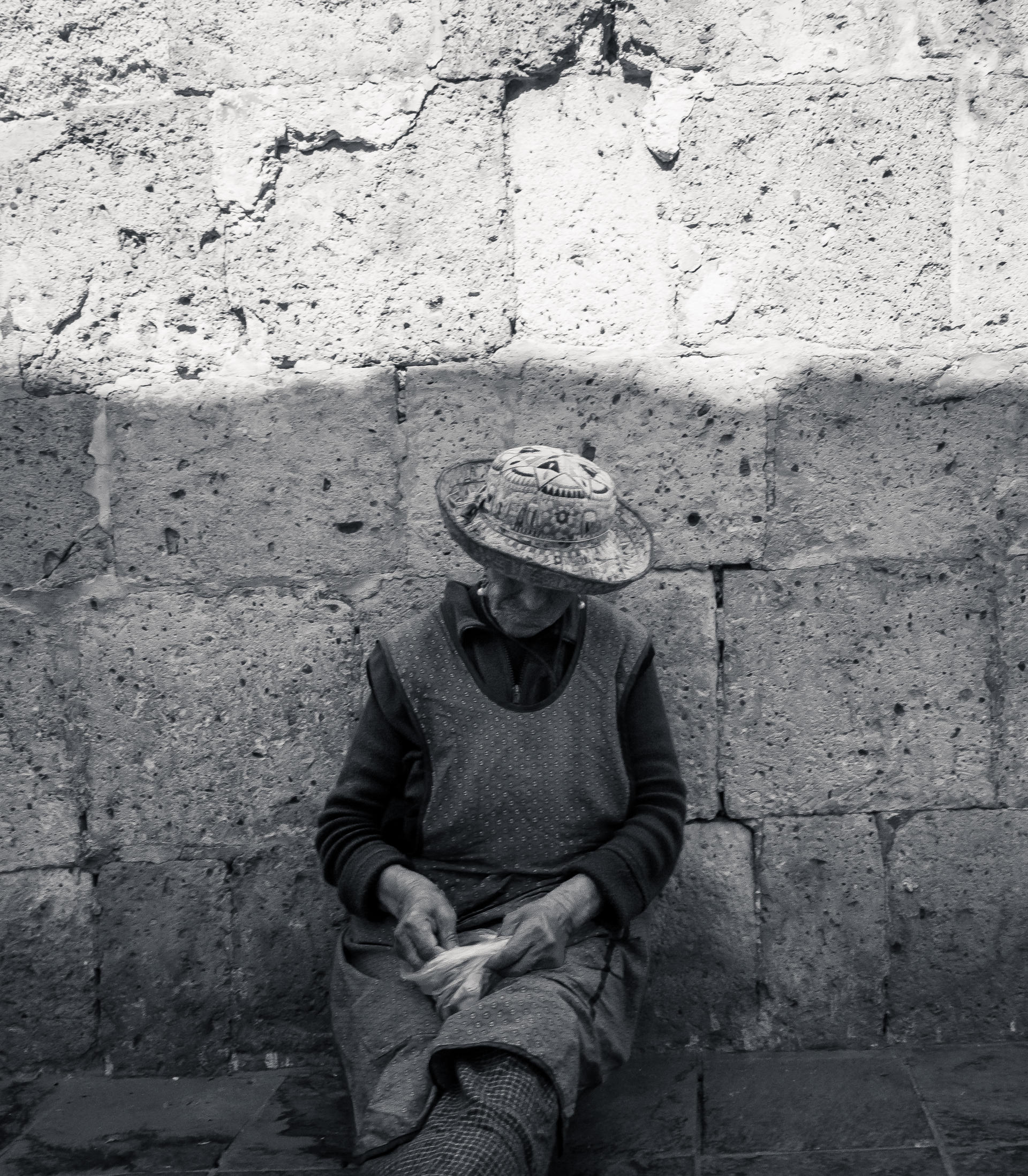 Old woman with hat sitting against stone wall, holding a small object.