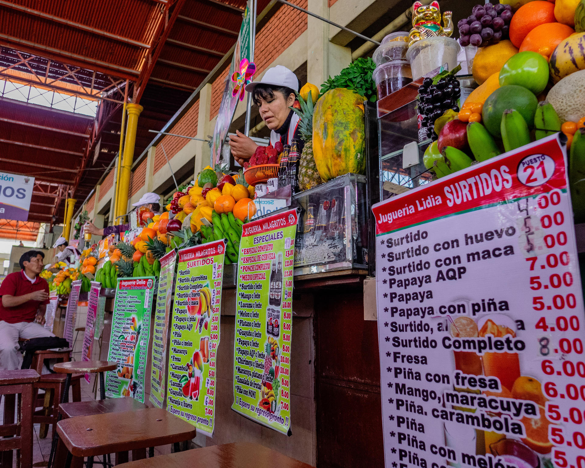 A vibrant market stall with fresh fruits, a chef preparing food, and colorful menu signs displaying prices.