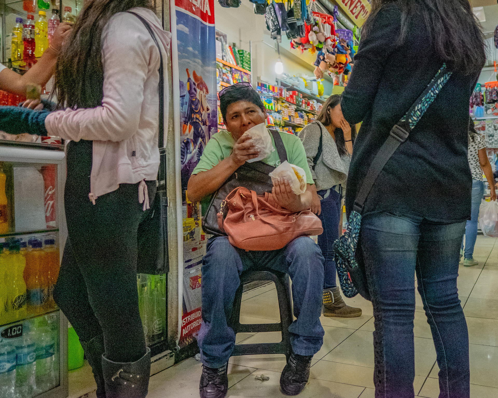 A man sitting on a stool in a store, eating from a bag and holding a drink.