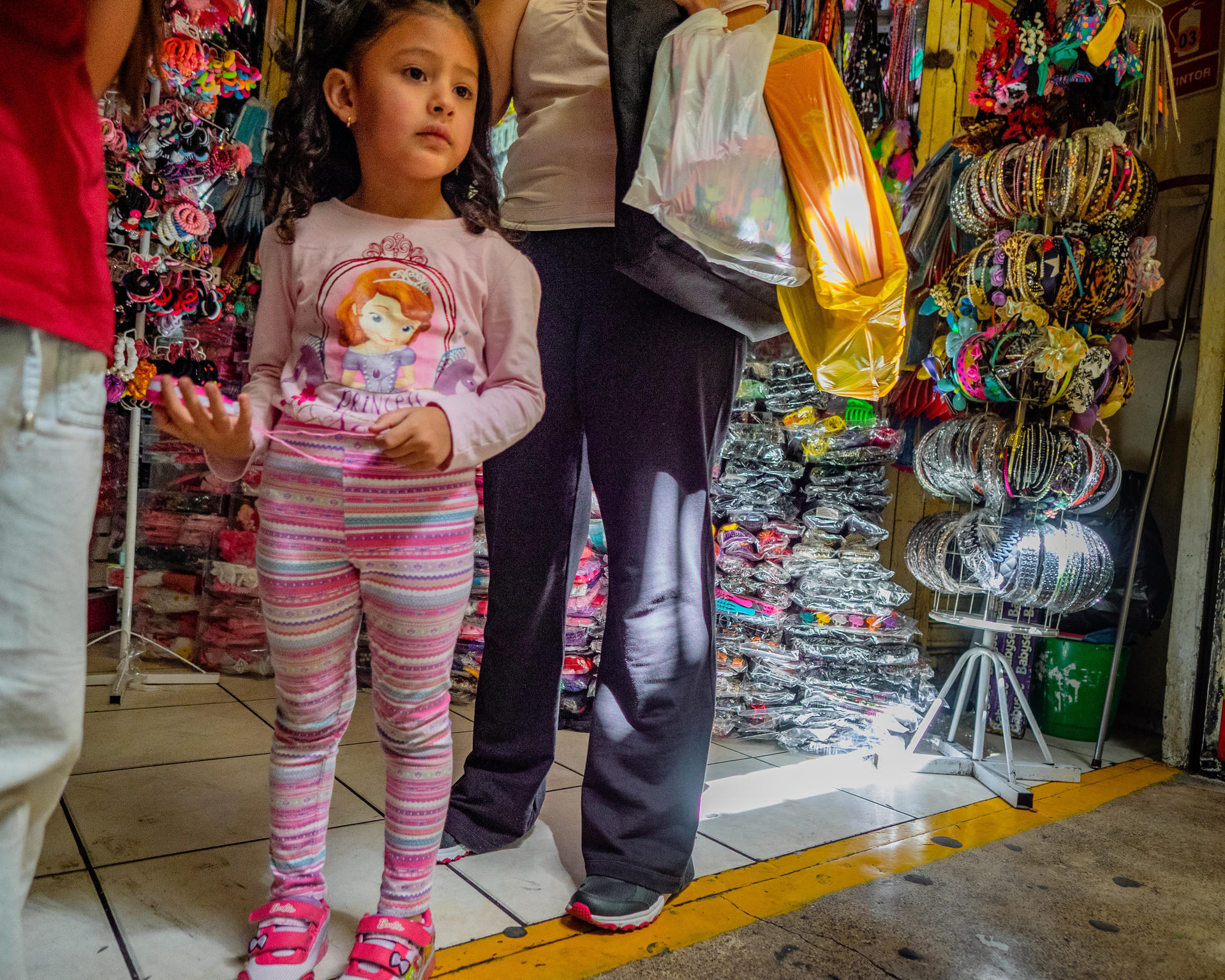 Little girl in pink striped pants and long-sleeve shirt, standing in a market with an adult.
