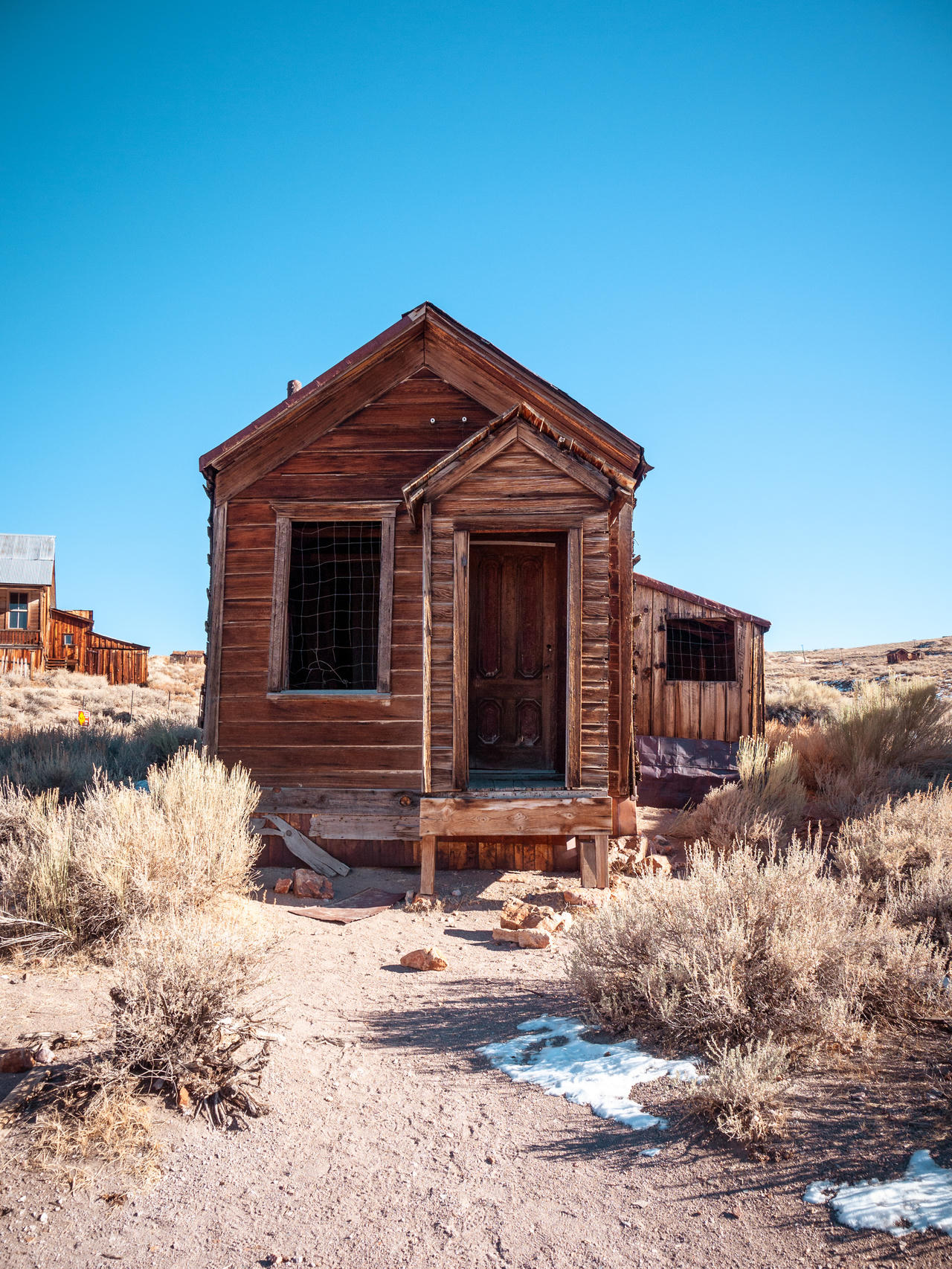 Abandoned wooden cabin in desert landscape with overgrown shrubs, clear sky