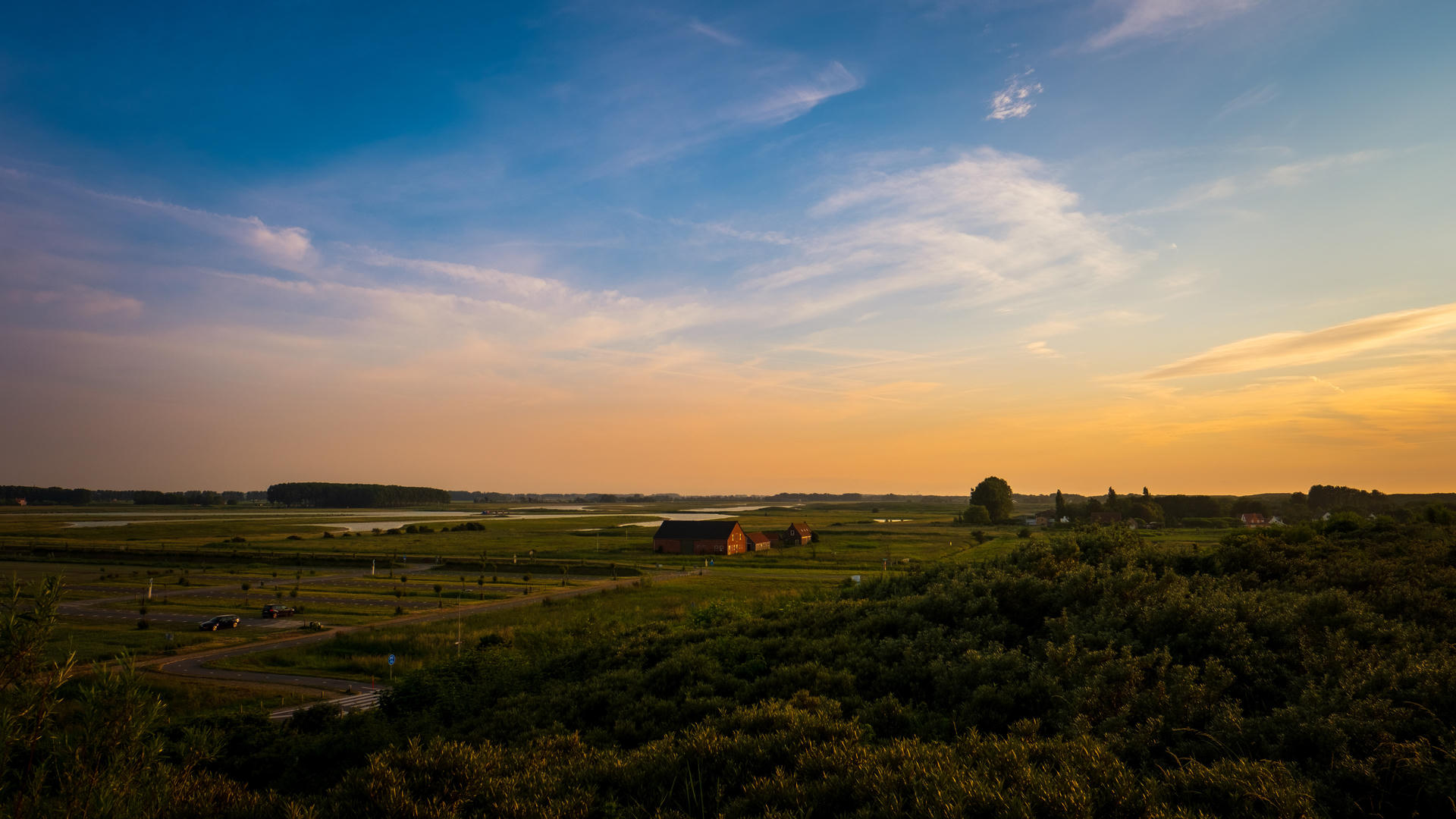 Sunset over green field with scattered trees and a barn on the horizon, sky transitioning from blue to orange.