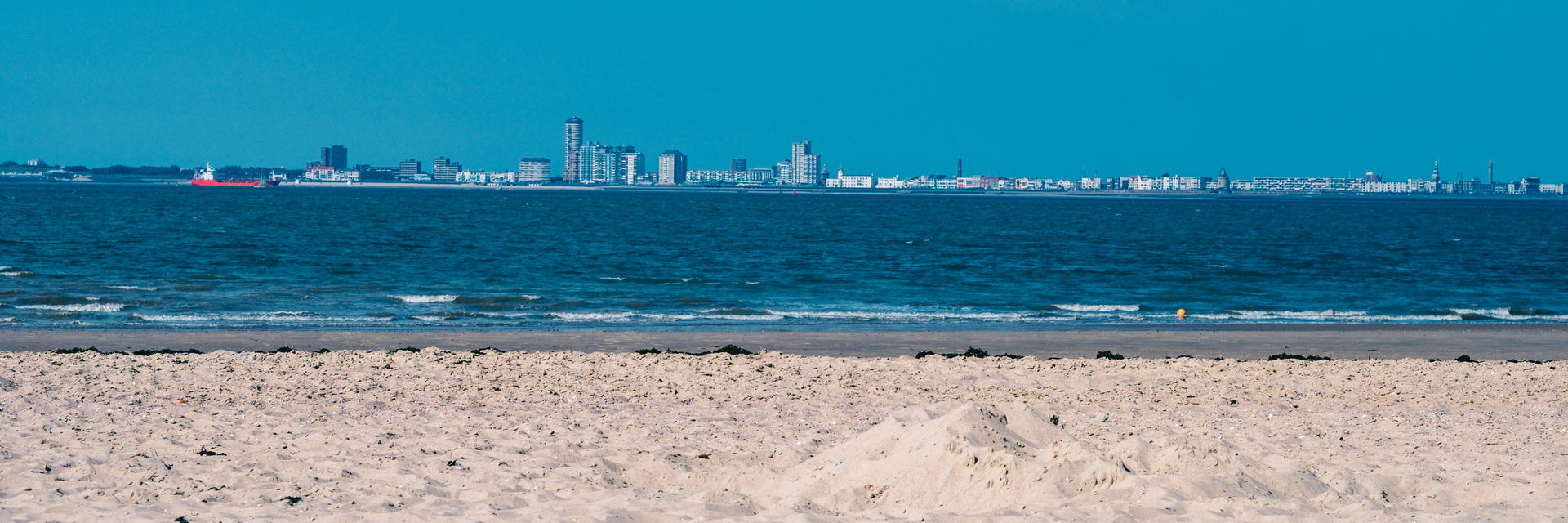 Panoramic view of an urban skyline across a calm blue sea, with a sandy beach in the foreground.