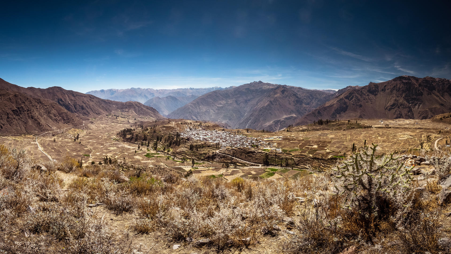 Desolate mountainous landscape with a small settlement nestled in the valley, sparse vegetation, and clear blue sky.