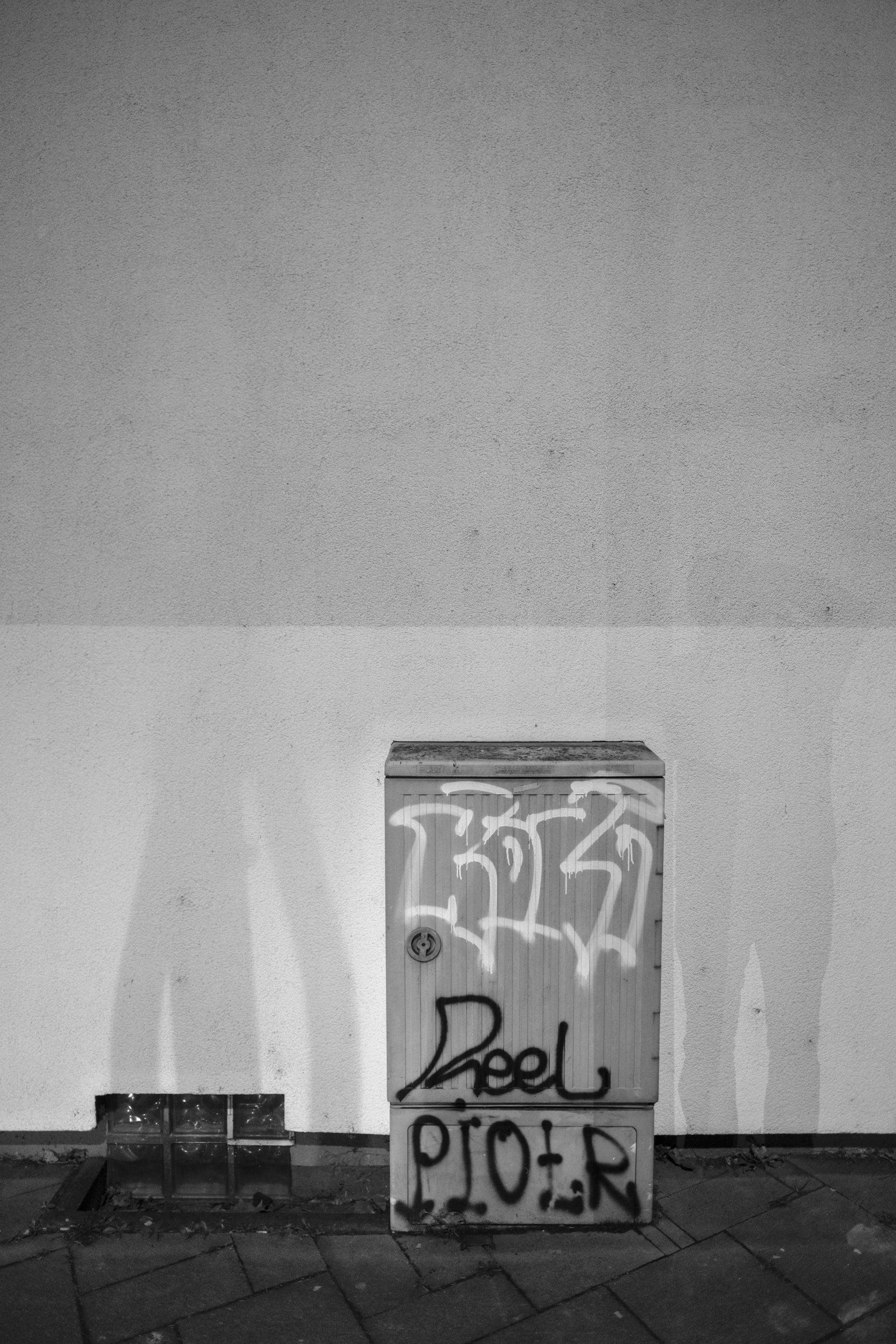 A graffiti-covered electrical box on a white wall with brick pavement, black and white.
