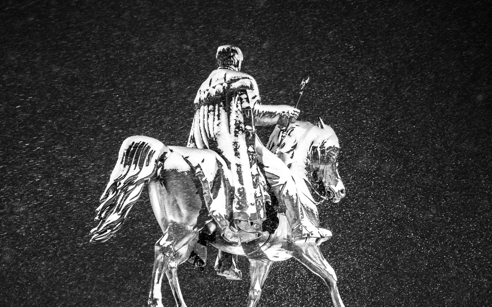 Man in a long coat riding a horse at night, rain falling around them.