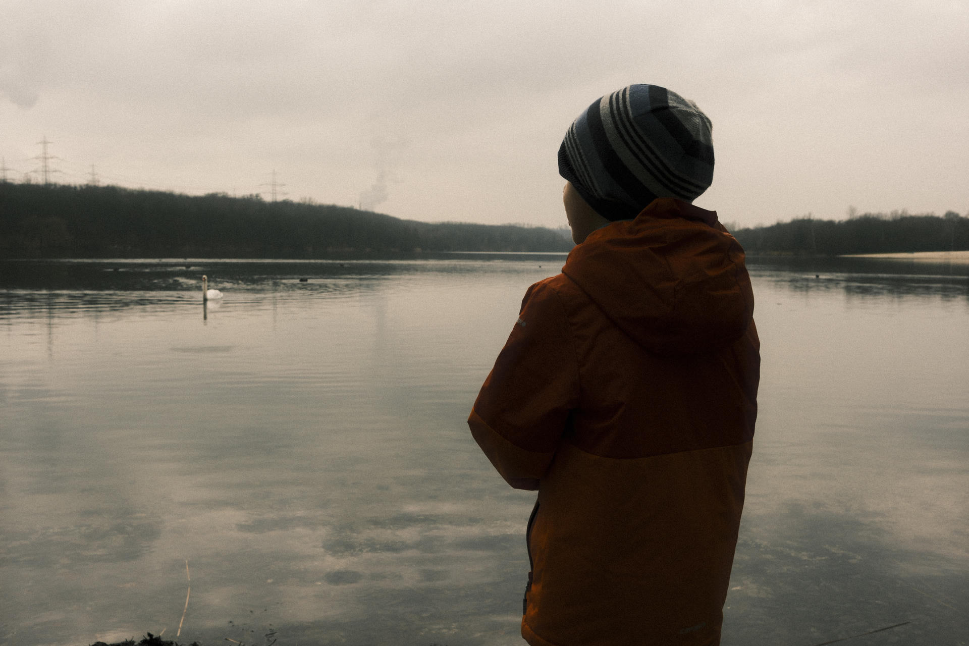 Person in orange jacket and gray beanie standing by calm lake, looking away from camera.
