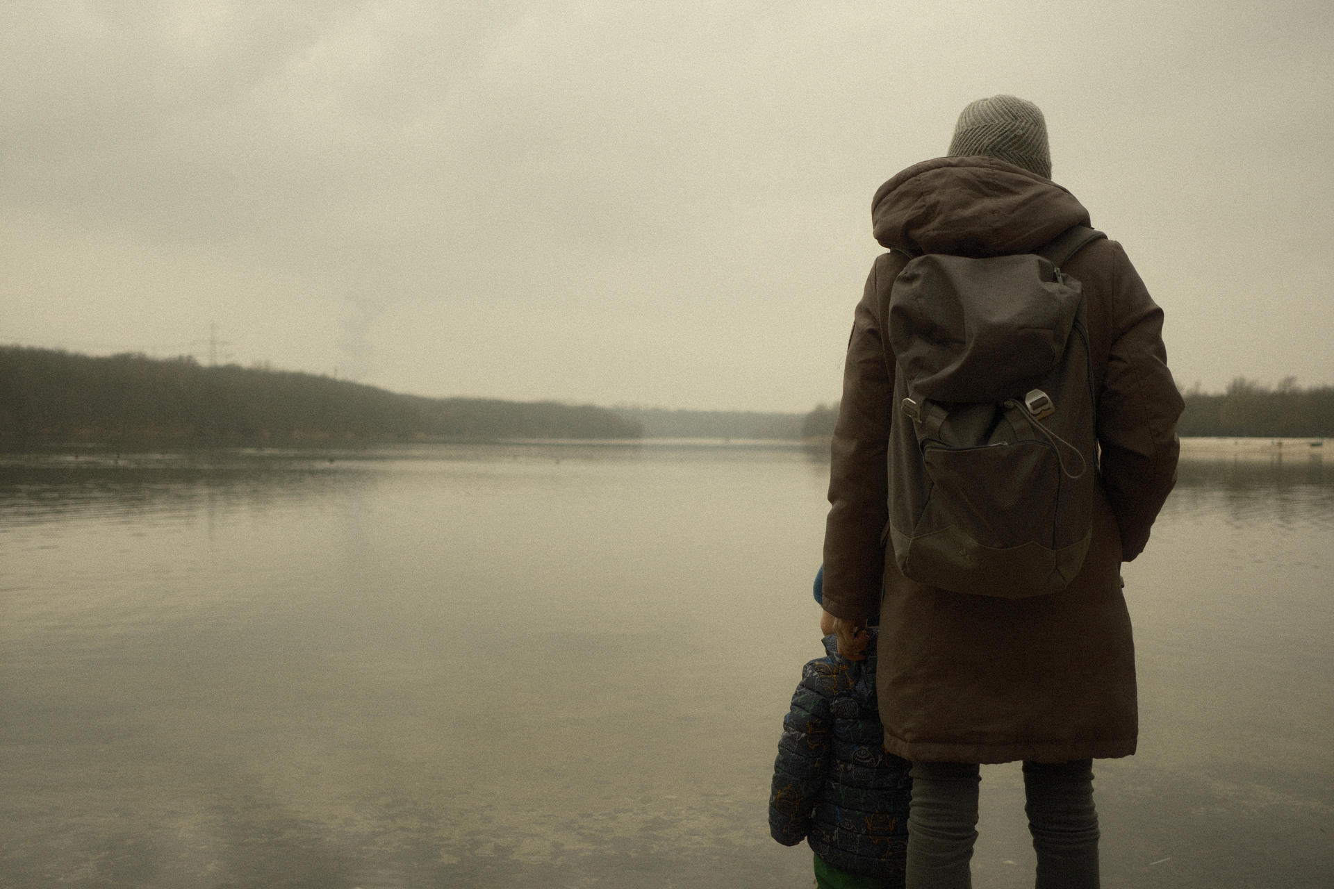 Person in coat and backpack standing by calm lake, foggy surroundings