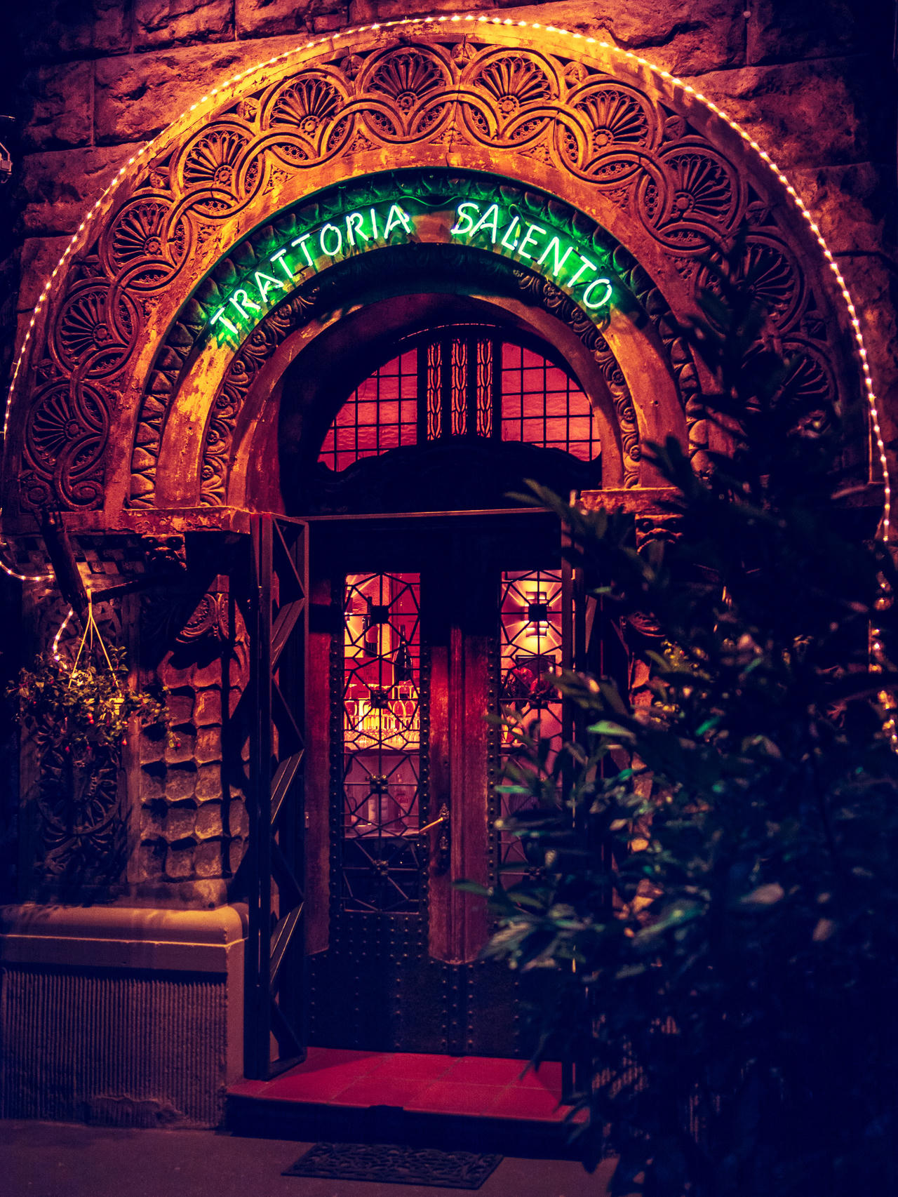 Restaurants entrance with ornate archway, neon sign, plants on either side, red carpet, and glass door.