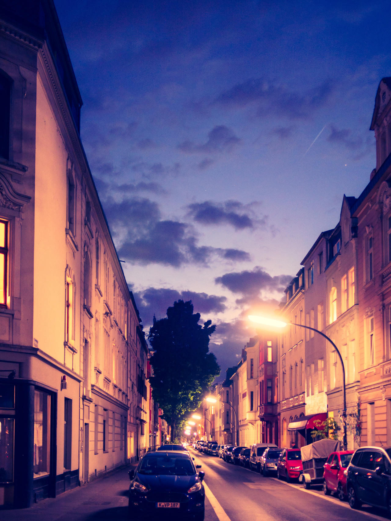A narrow street at dusk with buildings on either side, illuminated by a single streetlight.