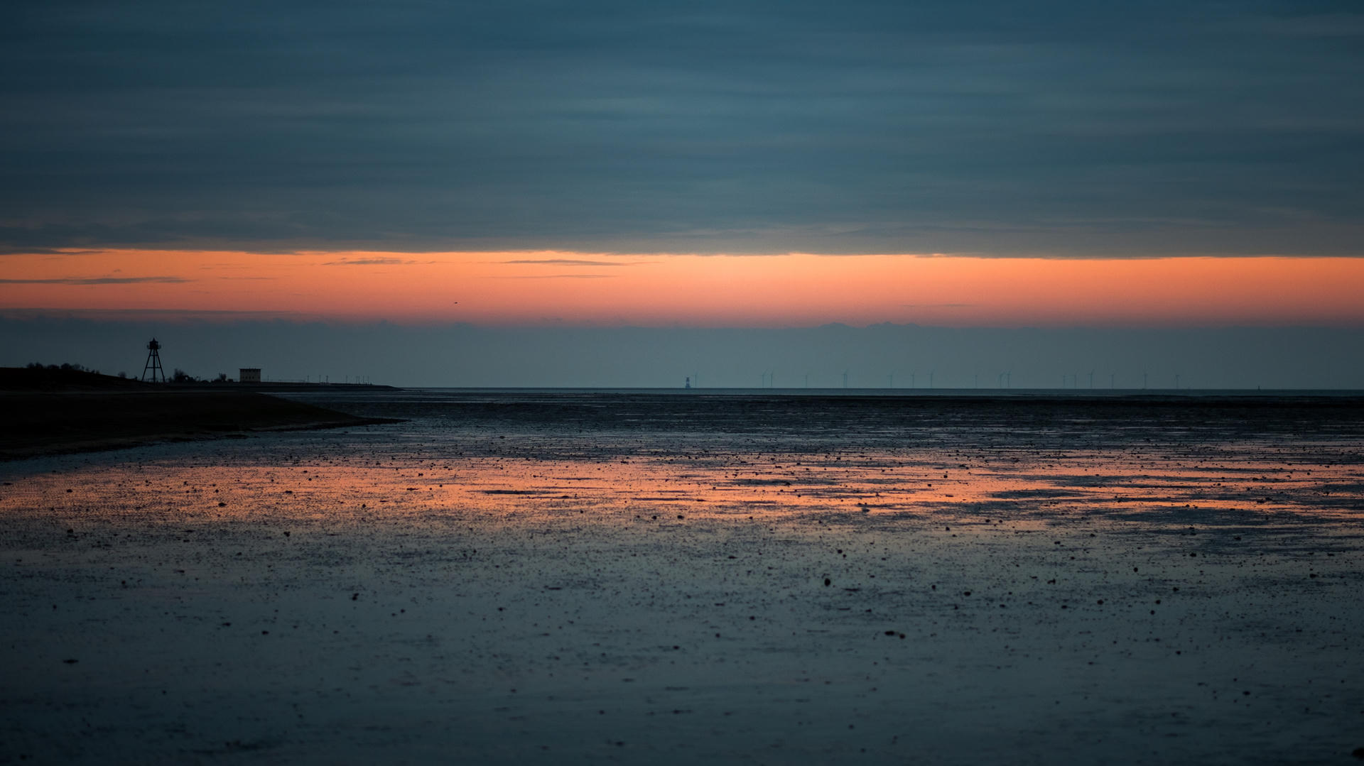 Sunset over calm beach with silhouetted lighthouse in background, vibrant sky reflected on wet sand.