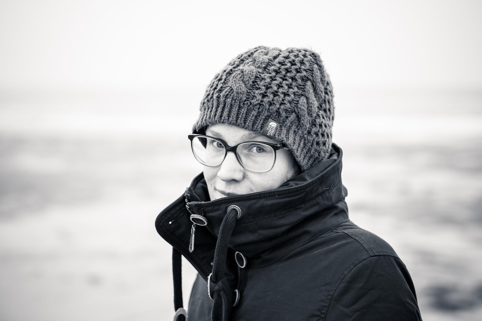 Person wearing glasses, knit beanie, and jacket, looking to the side against a blurred background.