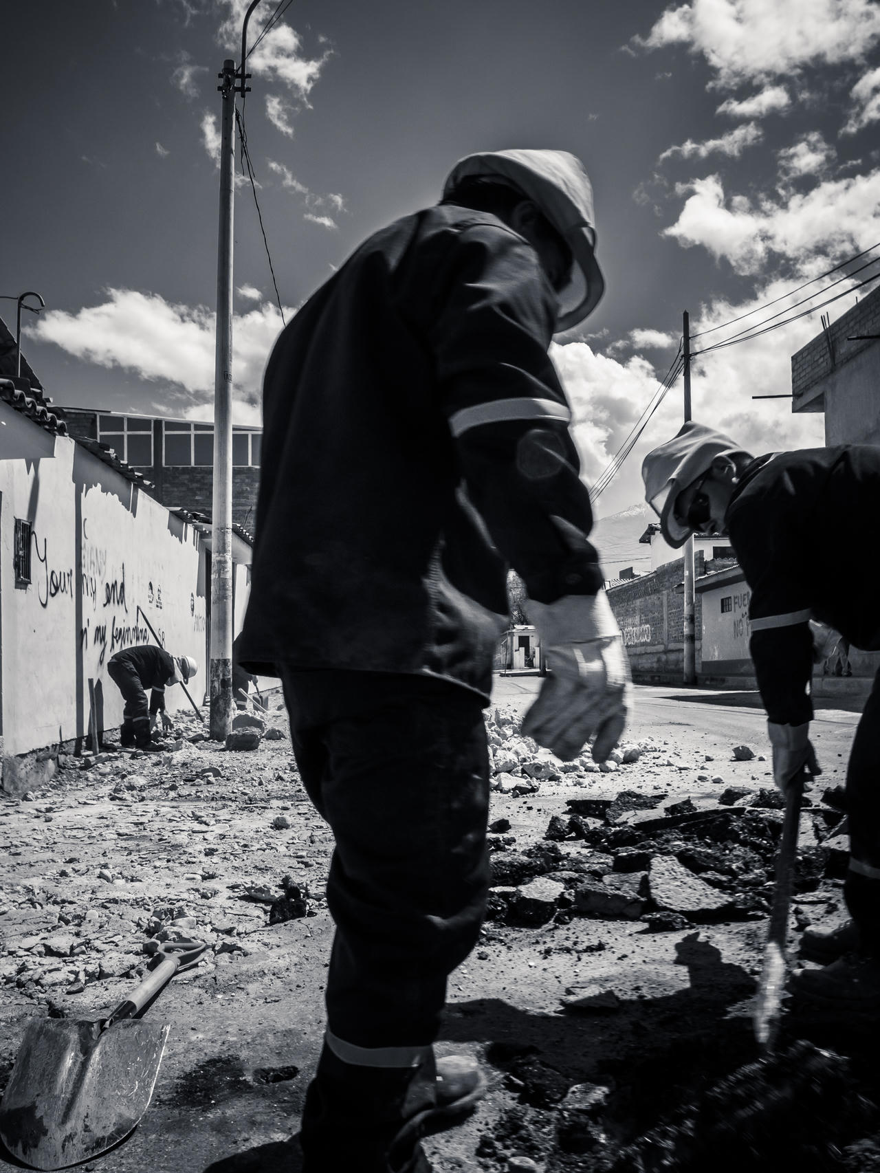 Three workers in helmets, two standing, one digging, debris on street, urban setting, black and white photo.