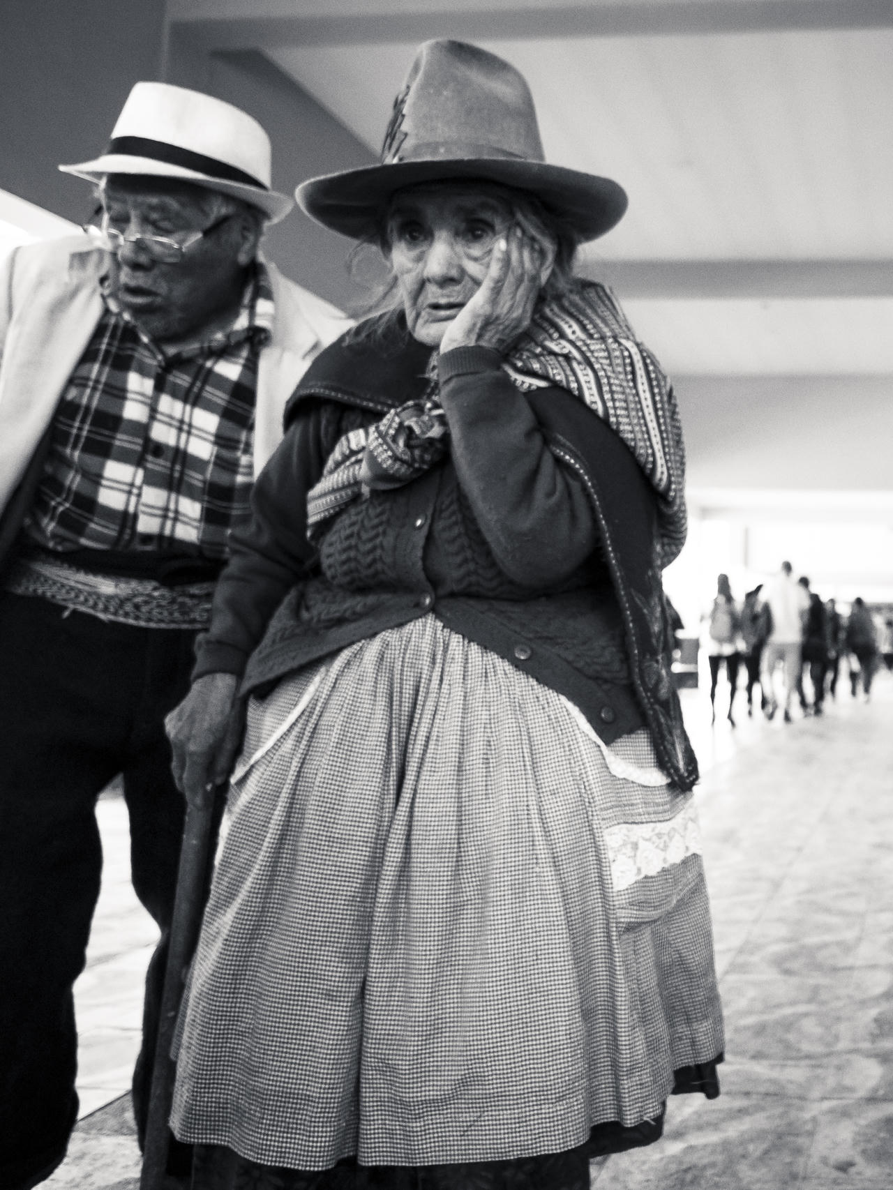 Woman in hat and plaid shawl, hand on face, man in hat and checkered scarf, blurred figures in background.