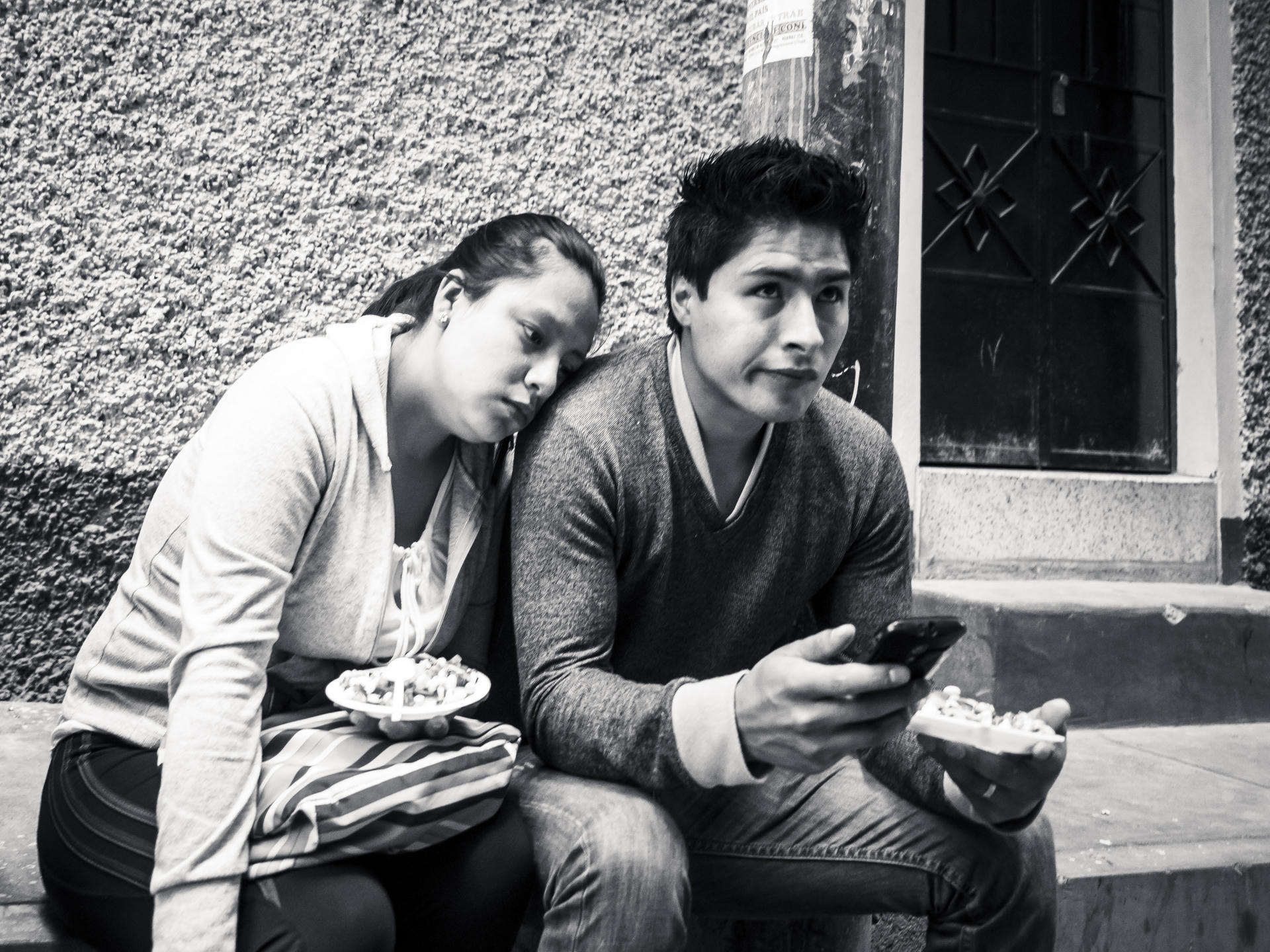 Couple sitting on steps, woman holding a bowl, man using smartphone, black and white.