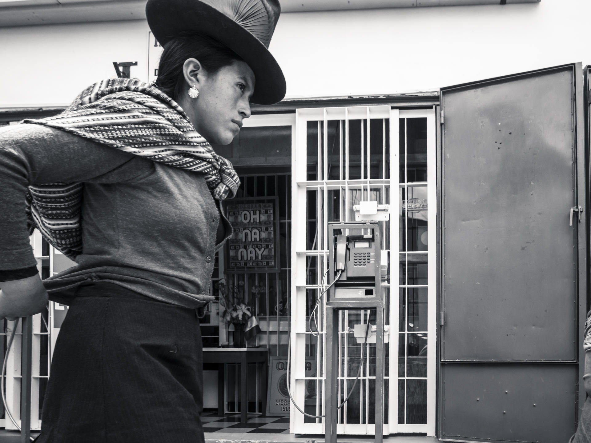 Person in traditional hat, scarf, looking away from a barred window with a telephone inside.