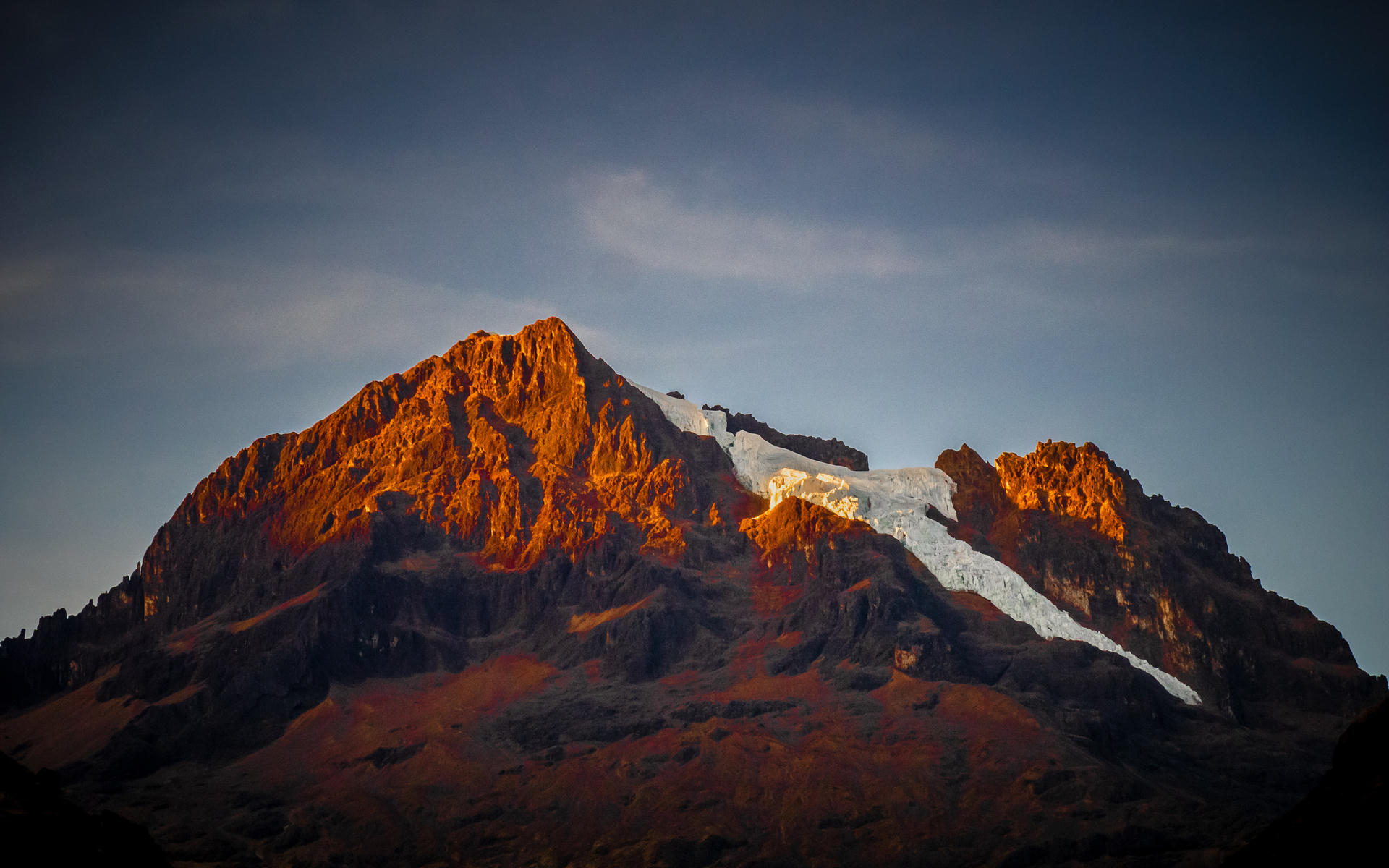 Mountain peak bathed in warm sunlight, with snow on its highest point against a cloudy sky backdrop.