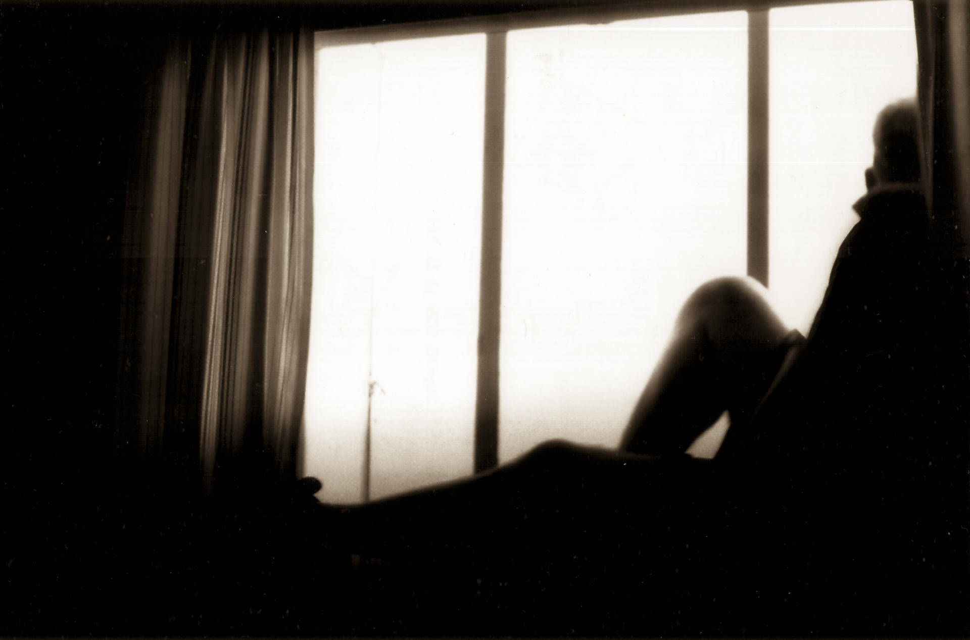 Person sitting on windowsill, backlit by bright light, silhouette against day-lit window, partially open curtains.