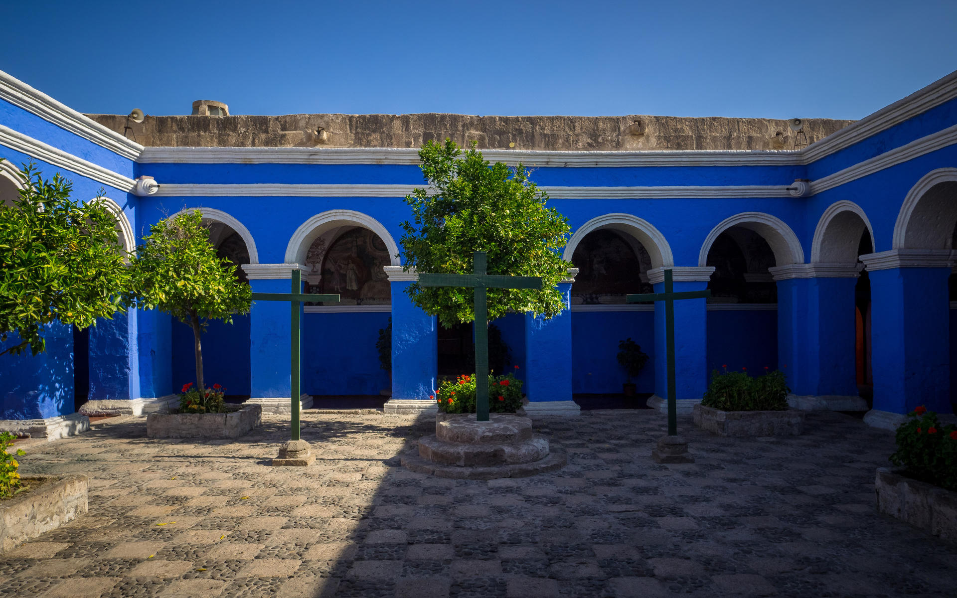 Bright blue courtyard with arches, potted plants, and a stone-tiled floor under clear sky.
