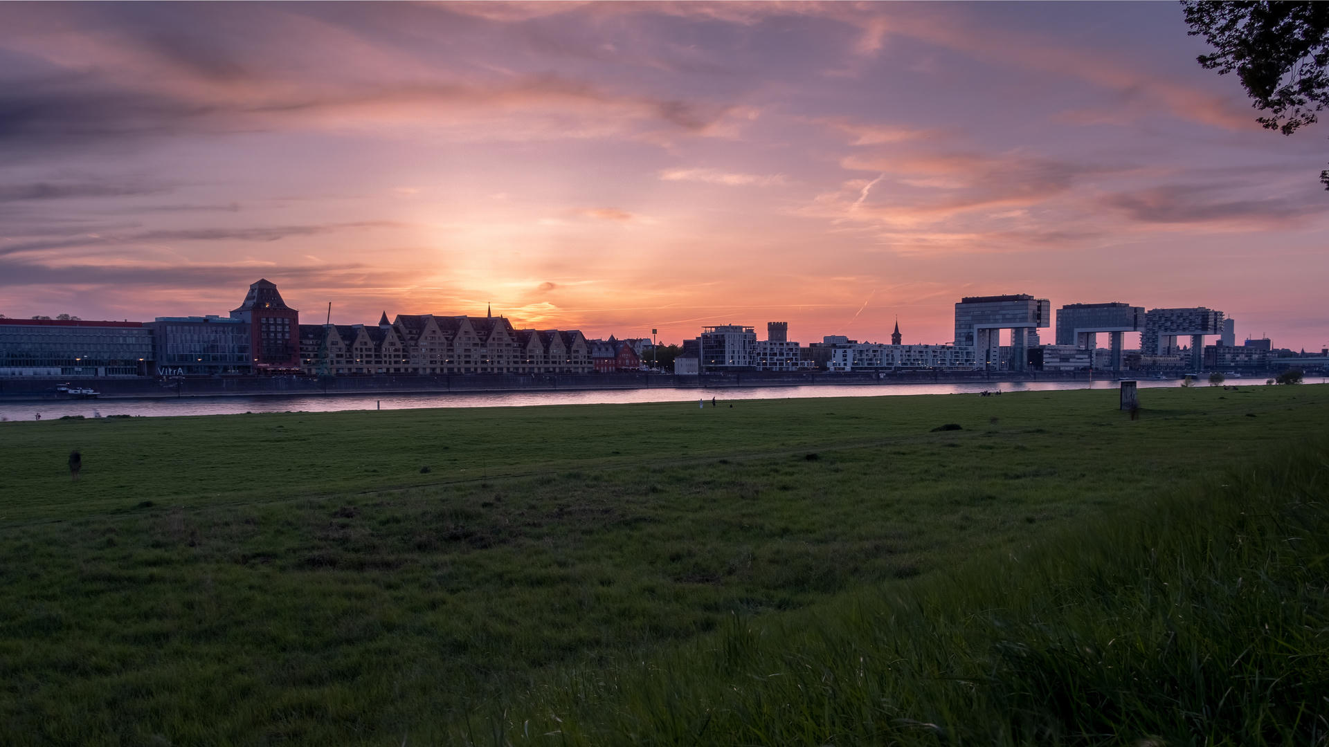 City skyline at sunset with buildings reflected in calm water, green field foreground.