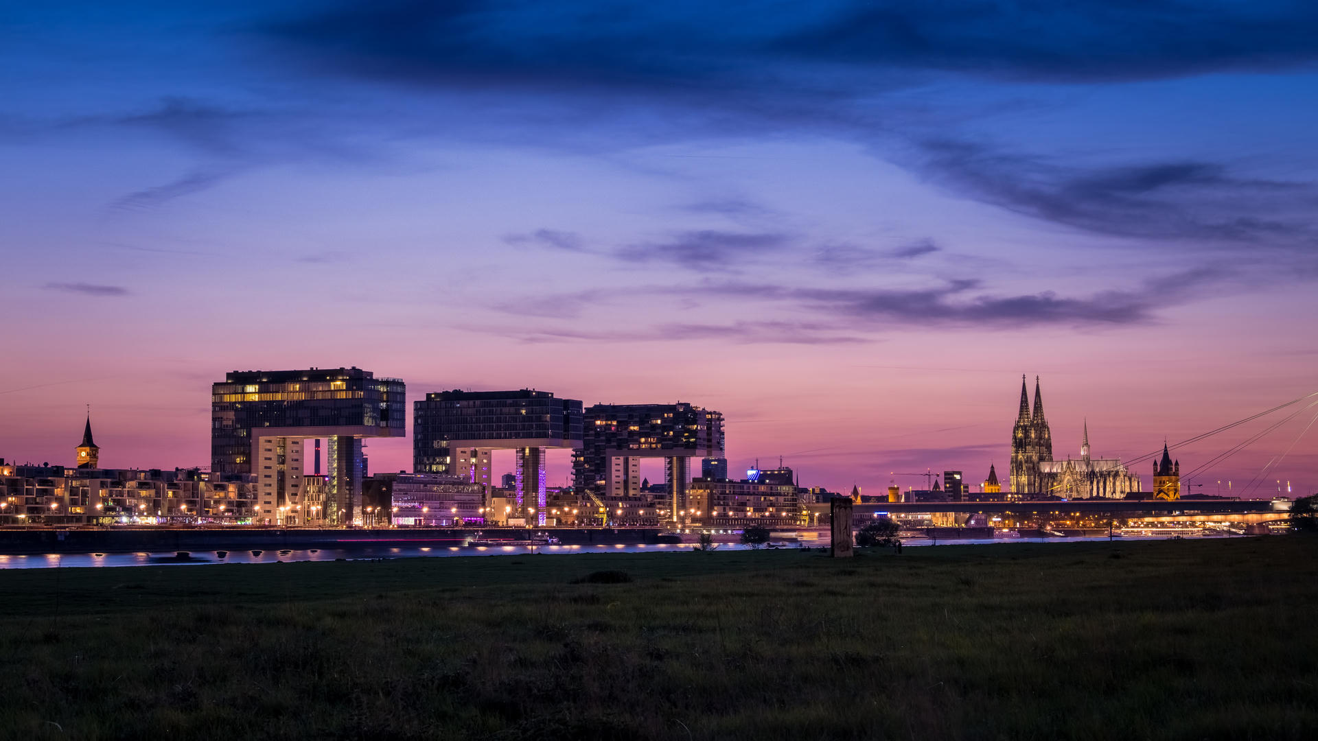 City skyline at dusk, buildings illuminated against a purple and blue sky with scattered clouds.