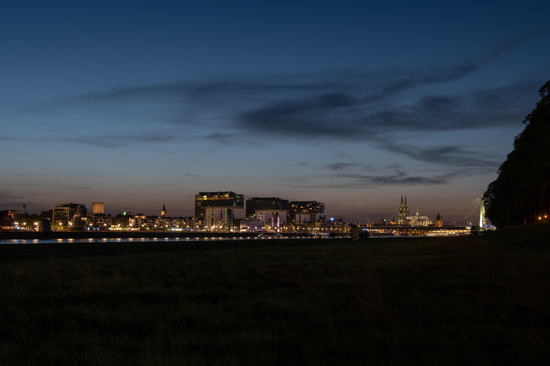 City skyline at dusk, buildings illuminated against dark blue sky with scattered clouds, water in foreground.