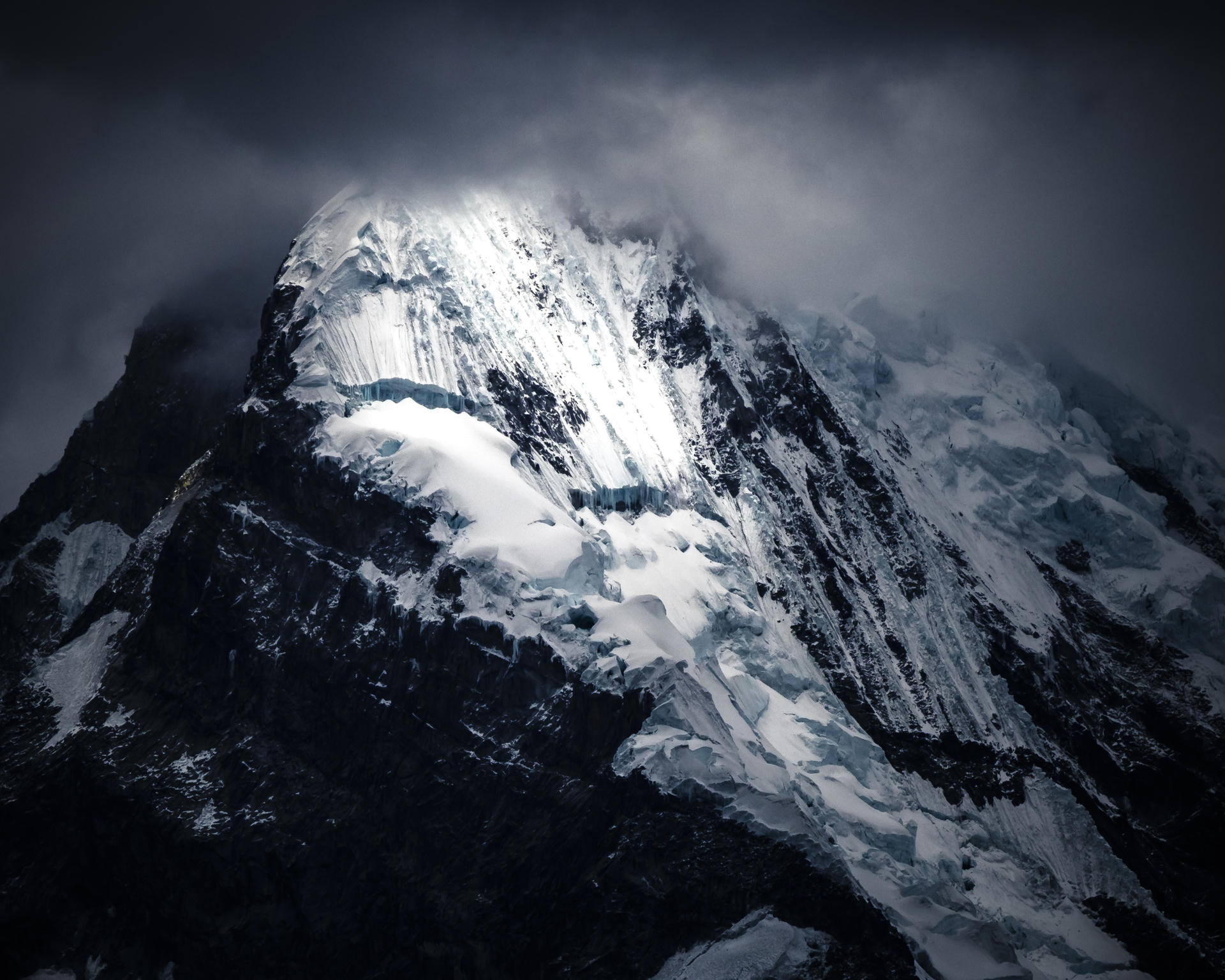 Snow-capped mountain peak emerging from dark clouds, light illuminating the summit.