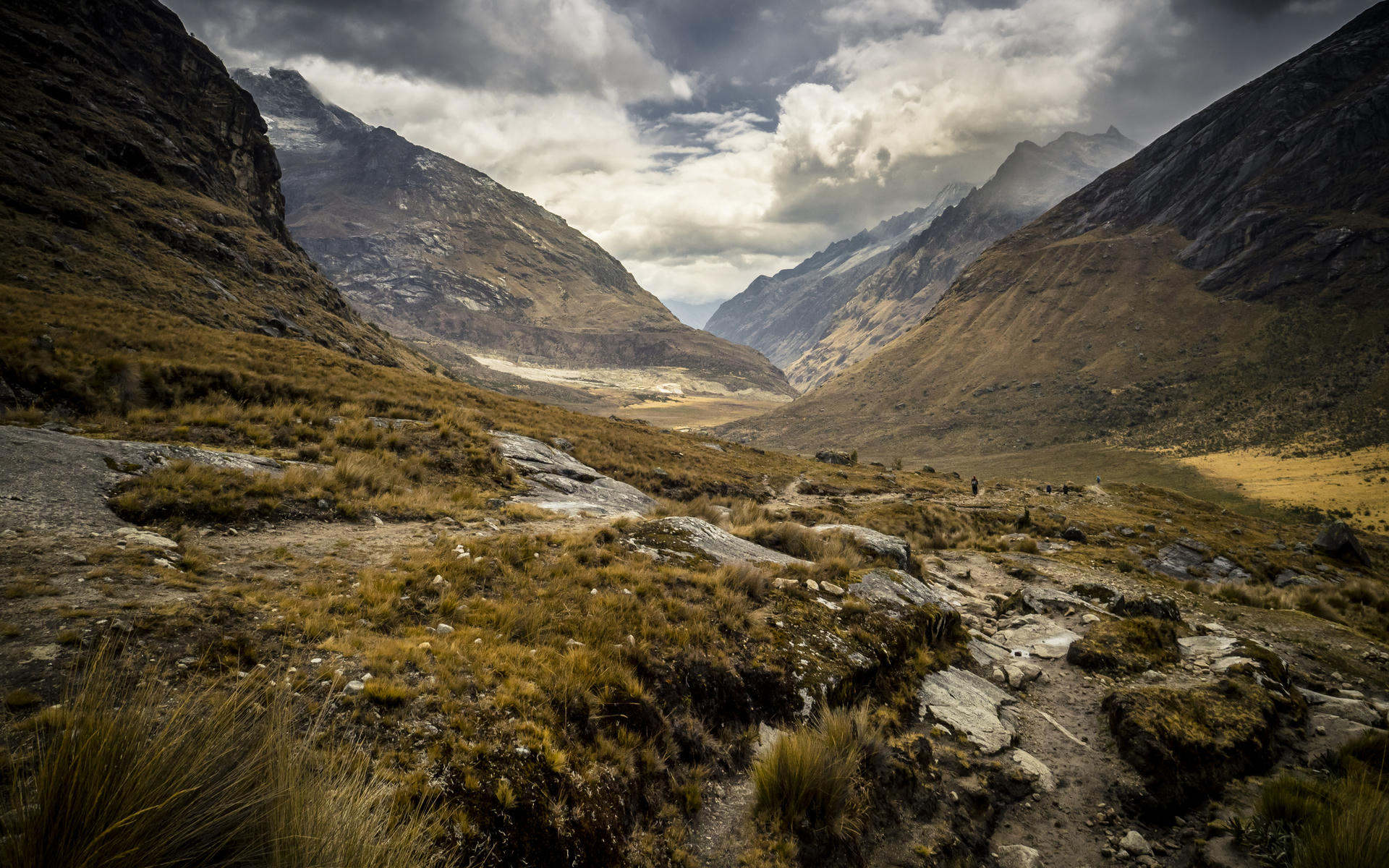 Mountain valley with rugged terrain, patchy grass, and dark clouds looming above.