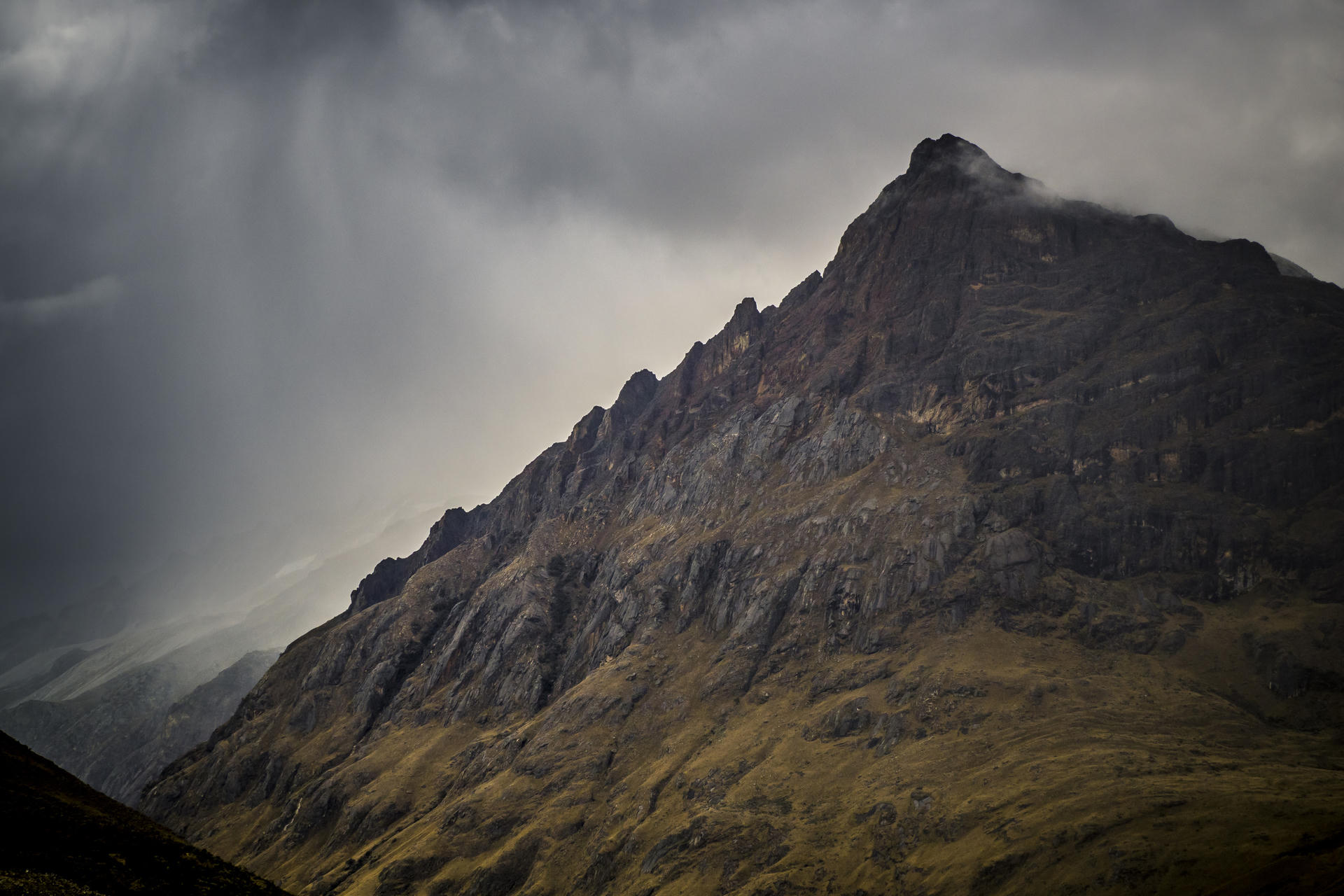 Dark, moody mountain peak with rocky face, set against dramatic cloud-filled sky.