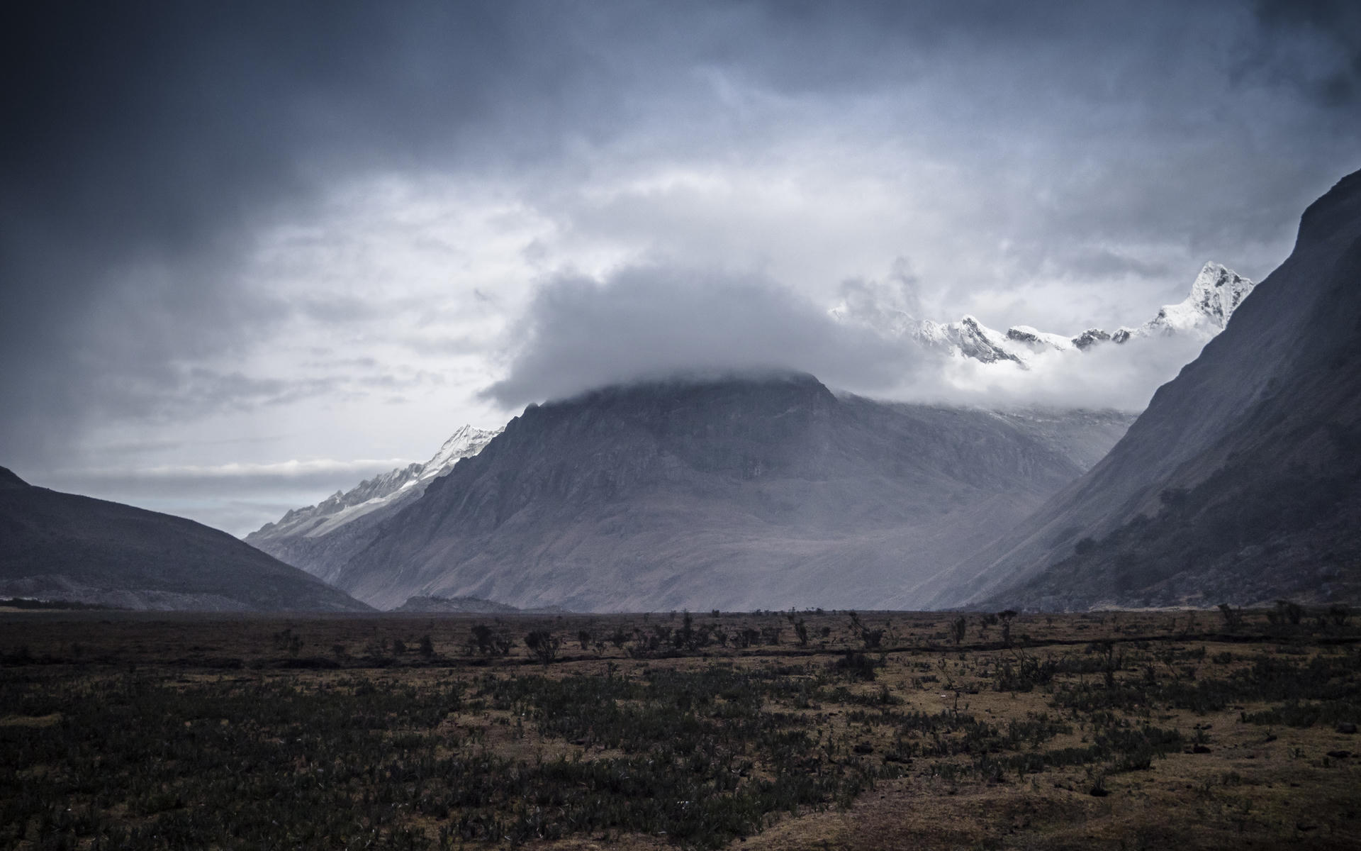 Mountain range with snow-capped peaks, partly obscured by dramatic clouds, set against a vast, barren landscape.