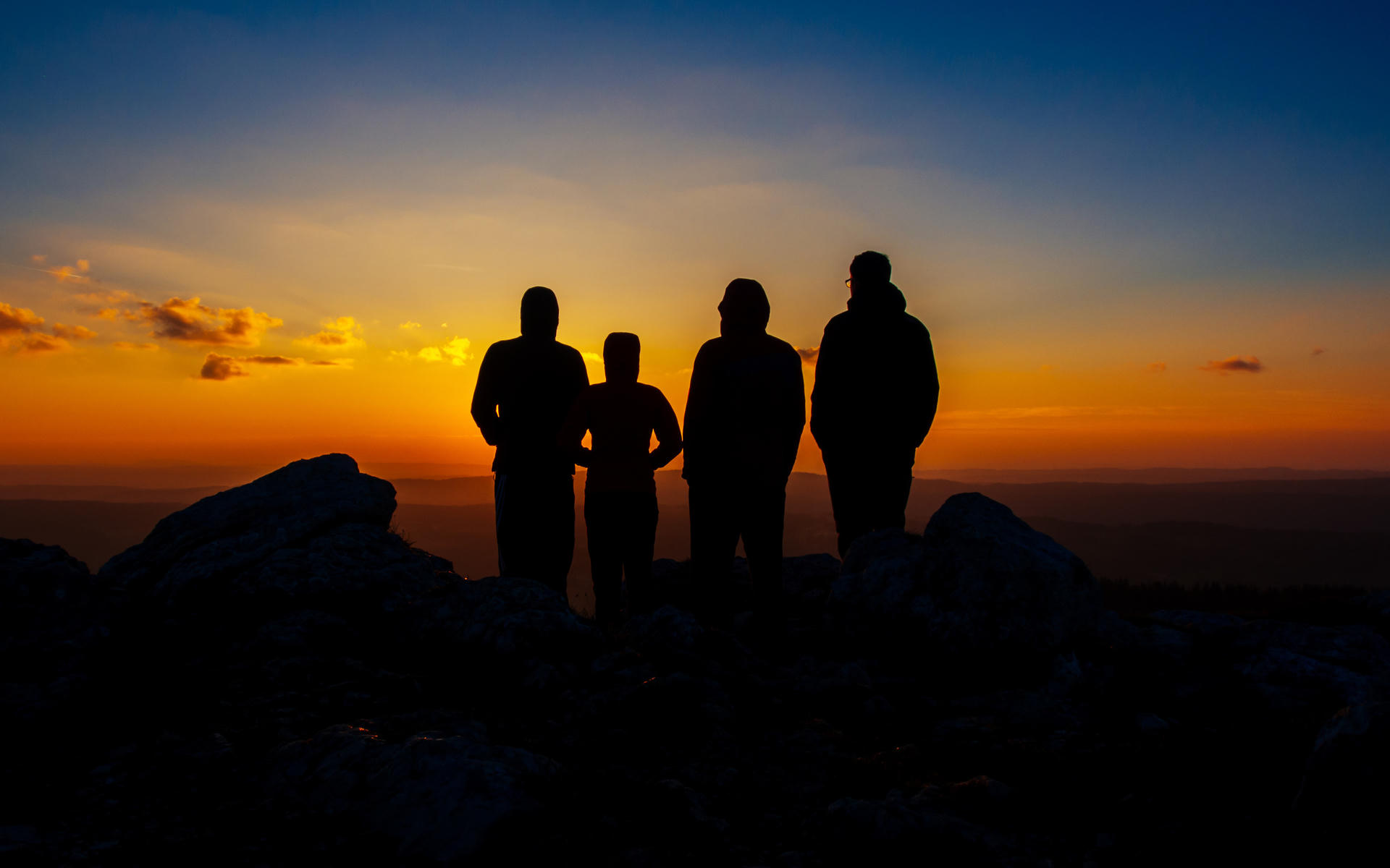 Four silhouetted figures standing on rocky terrain at sunset, facing a vibrant sky transitioning from orange to blue.