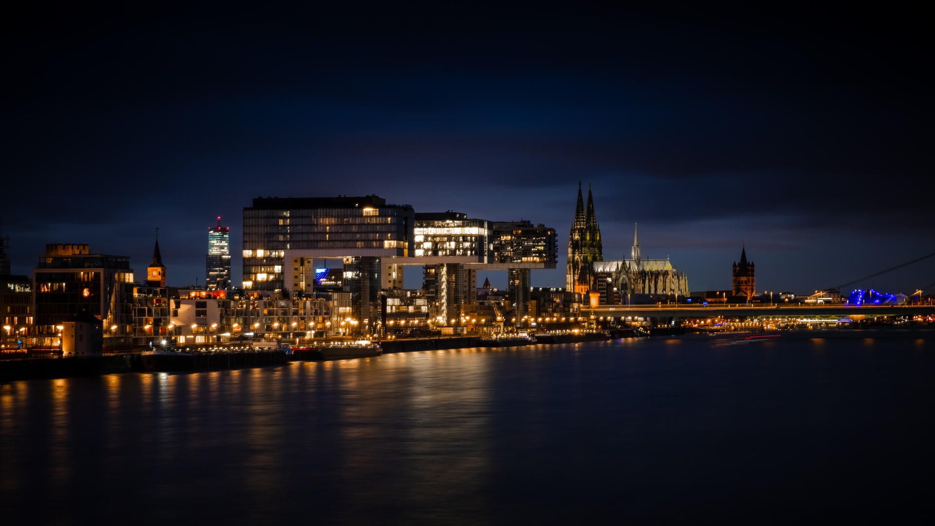 City skyline at night with illuminated buildings reflected in calm water, a bridge on the right side.