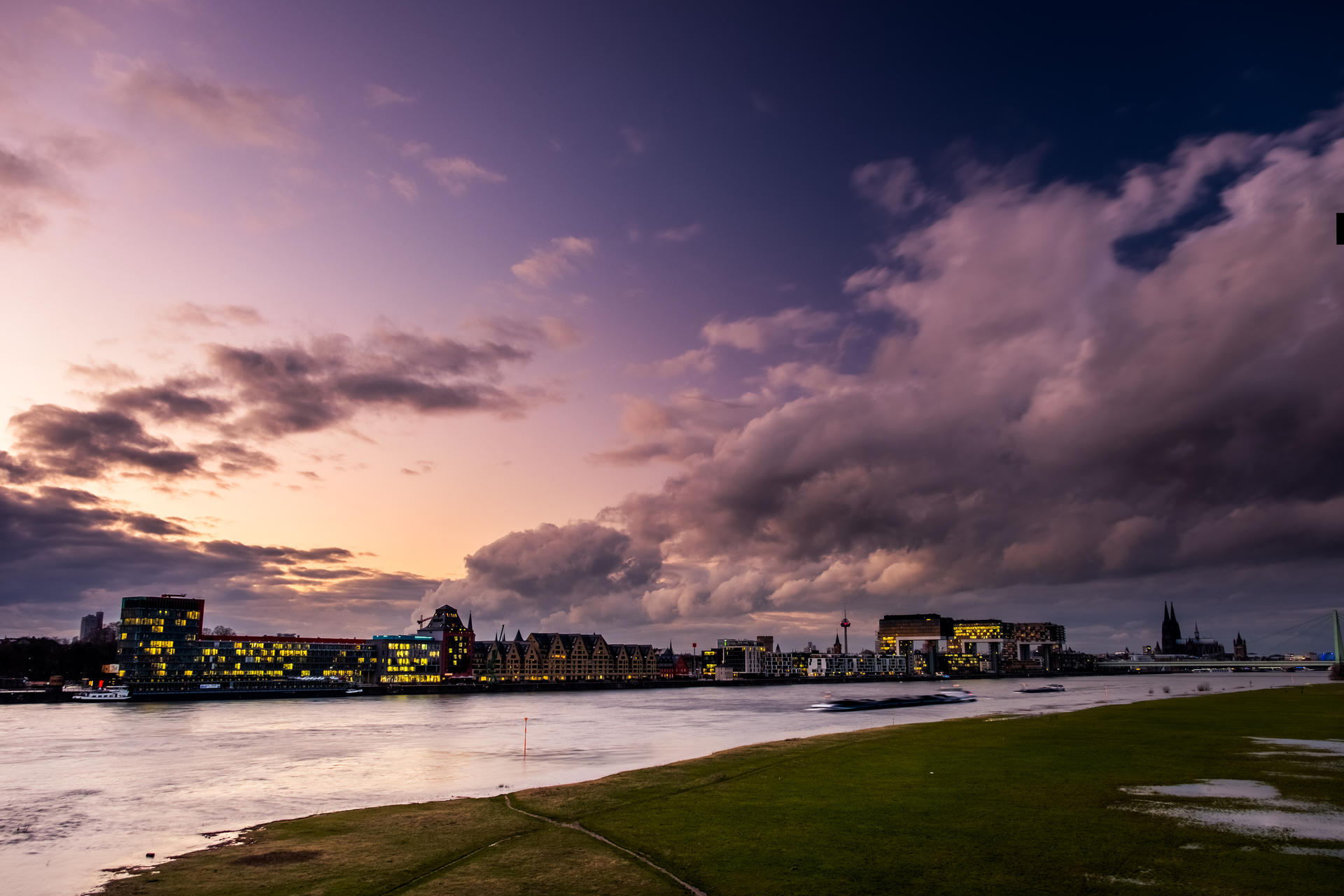 Sunset over city skyline with illuminated buildings, clouds, and a grassy foreground.
