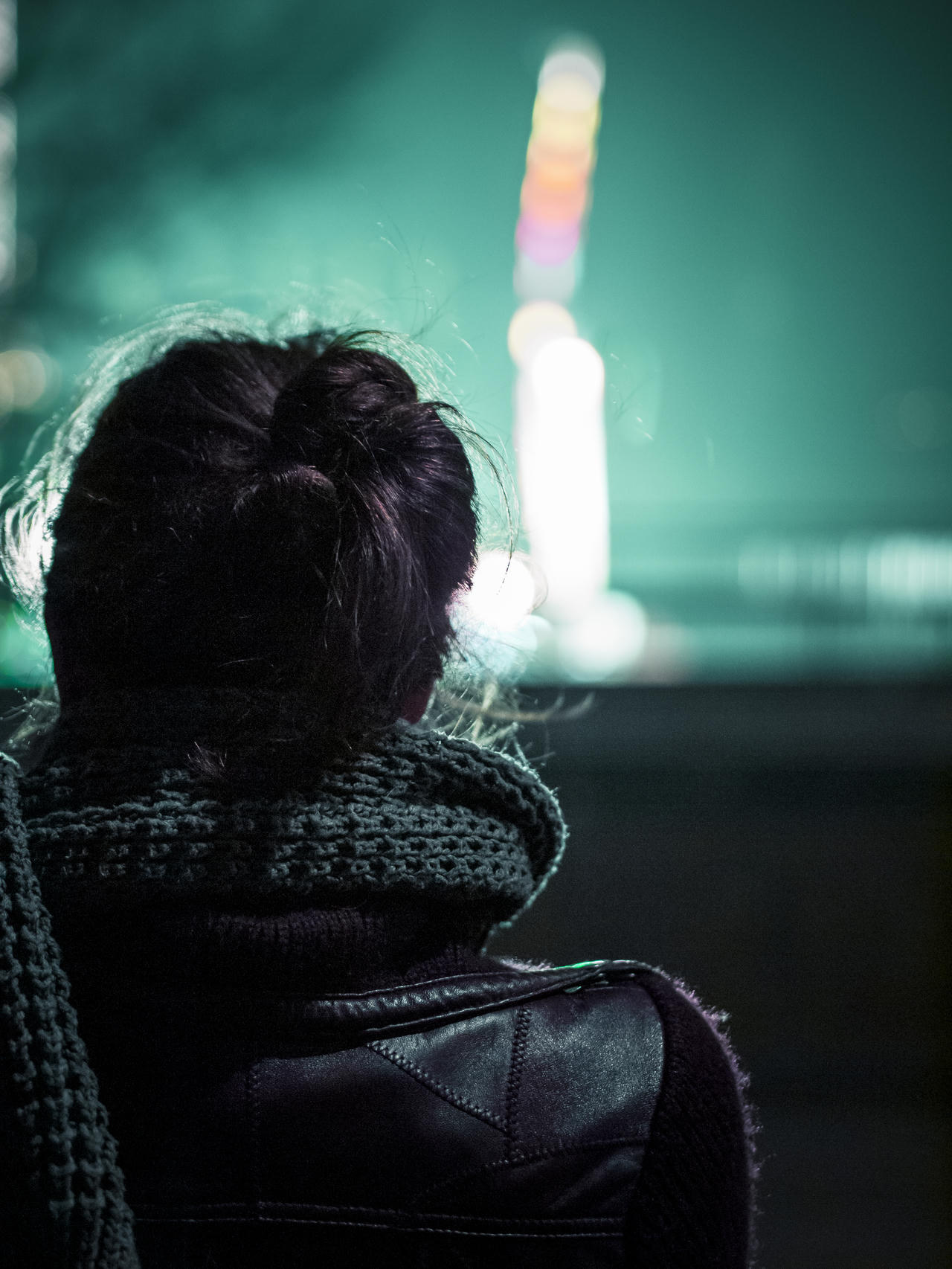 Person with dark hair in bun, wearing jacket, facing away from camera at night with blurred lights background.