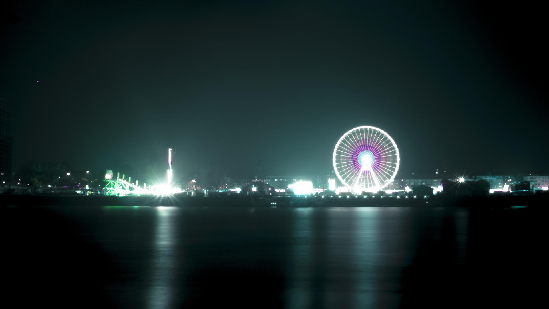 Night view of illuminated Ferris wheel and city skyline reflected on calm water, with a lit tower in the distance.