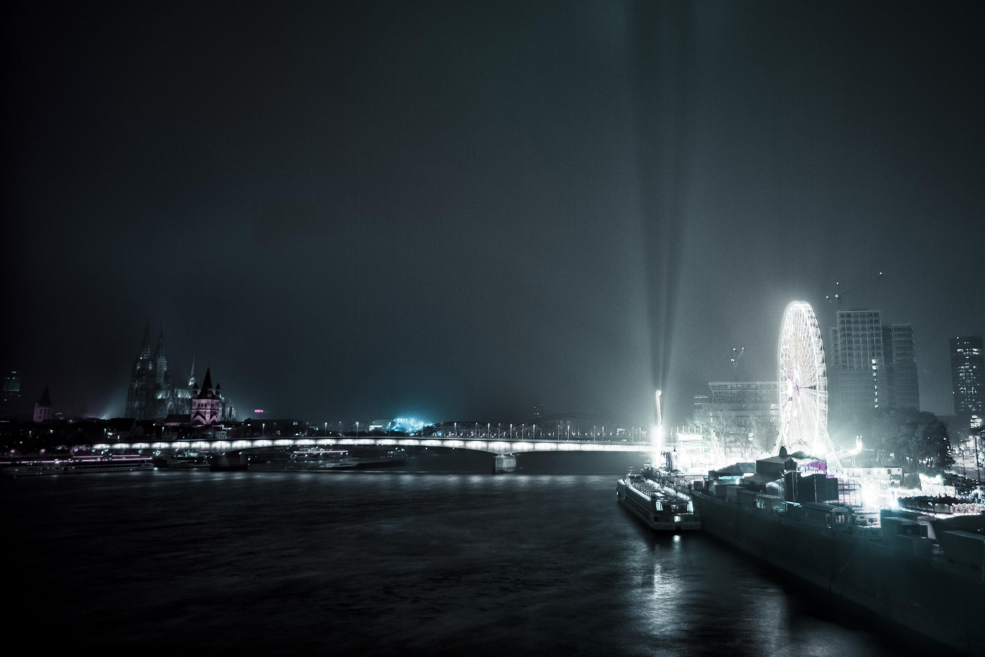 Nighttime cityscape with illuminated Ferris wheel, buildings, and searchlight beams over a river.